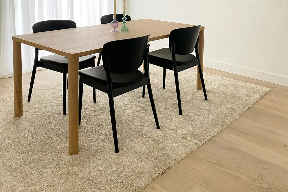 Een houten eettafel met vier zwarte stoelen op een licht vloerkleed, met vitrage voor het raam