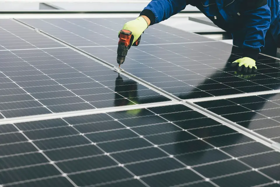 Een monteur in werkkleding schroeft met een accuboormachine een zonnepaneel vast aan de rail, tussen andere zwarte panelen