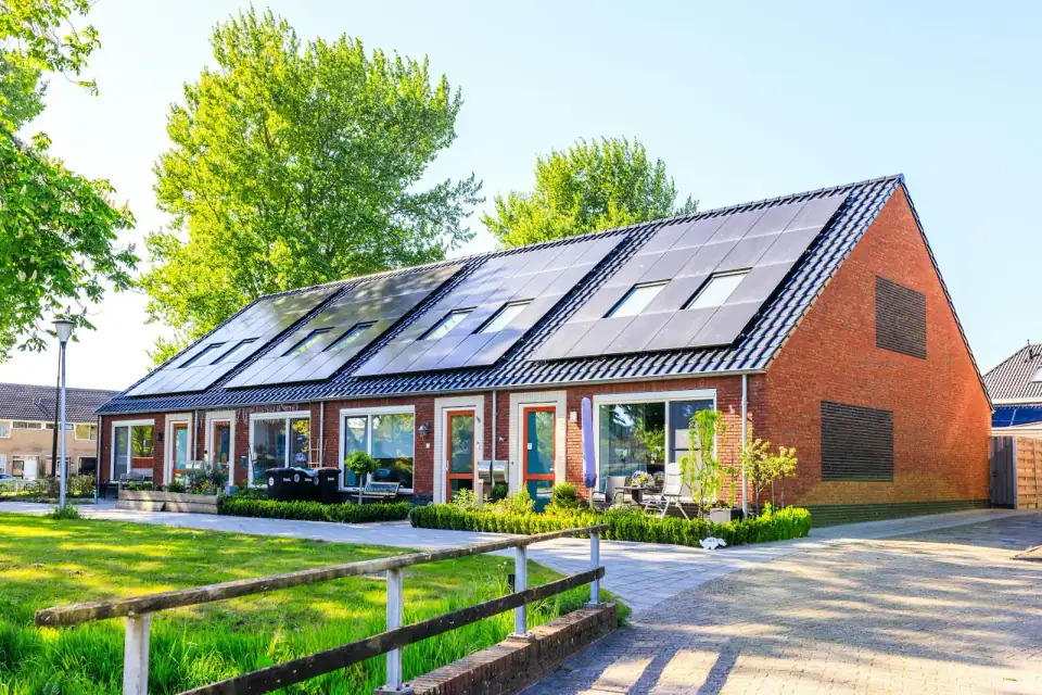 Een rij bakstenen woningen met zwarte zonnepanelen en dakramen op het dak, met voortuinen en een houten hek in de zon